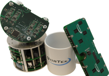 A white mug with the QorTek logo in between two green digital power amplifiers not much larger than the mug.