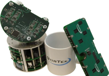 A white mug with the QorTek logo in between two green digital power amplifiers not much larger than the mug.